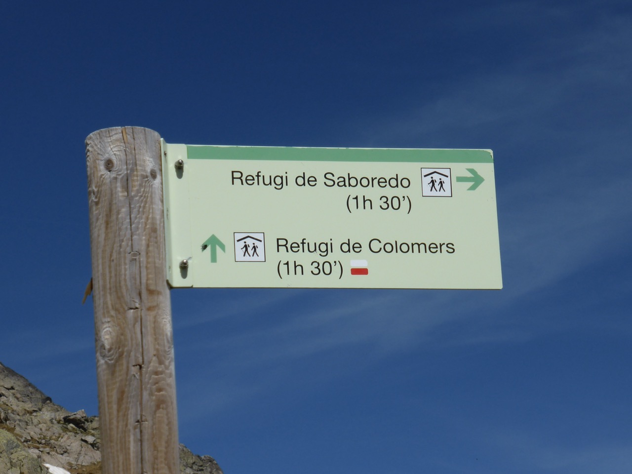 A wooden trail sign against a deep blue sky showing directions to Refugi de Saboredo (1h 30') and Refugi de Colomers (1h 30'), with the yellow-and-white flag marker