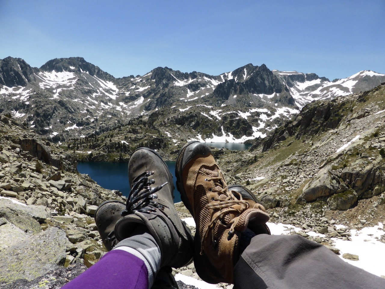 Two pairs of well-worn hiking boots resting on a rock in the foreground, with a panorama of alpine lakes and snow-patched jagged peaks stretching into the distance